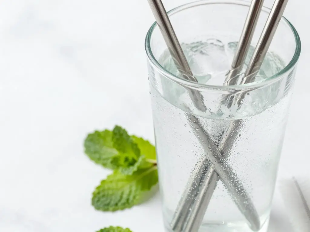 Artistic close-up photography of stainless steel straws in clear glass filled with iced beverage, condensation on glass, scattered fresh min.