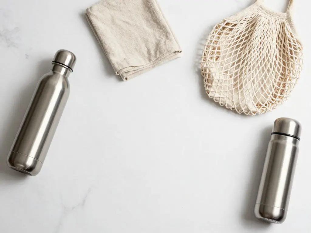 flat lay composition on a clean white marble countertop featuring essential reusable items: stainless steel water bottle, folded cloth napki.