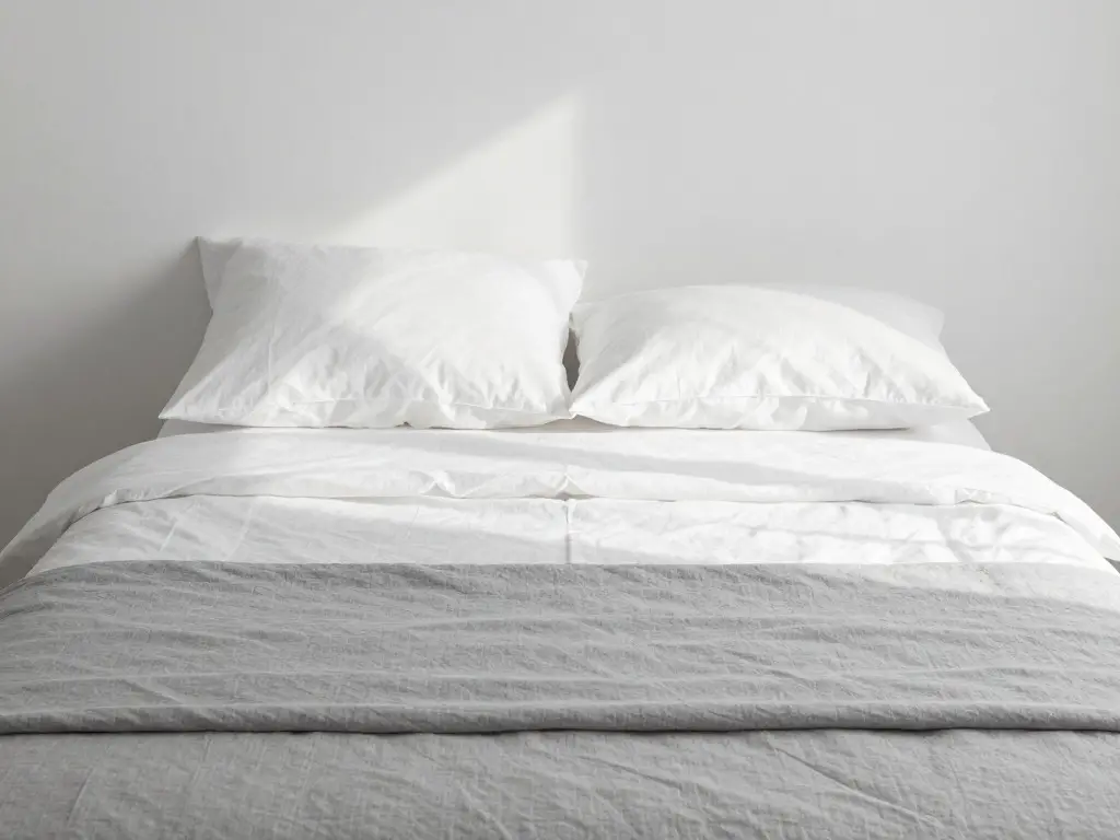 Neatly made minimalist bed with crisp white linen sheets and a simple gray comforter, morning sunlight casting soft shadows, clean and