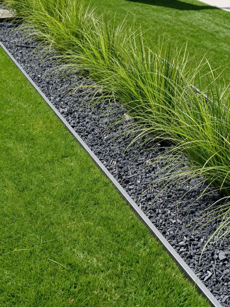 A clean, modern garden bed edged with sleek metal bordering separating vibrant green lawn from a minimalist planting strip filled with ornam.