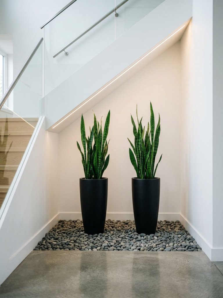 An ultra-minimal small garden under stairs featuring two tall black planters placed symmetrically on a bed of smooth grey pebbles.